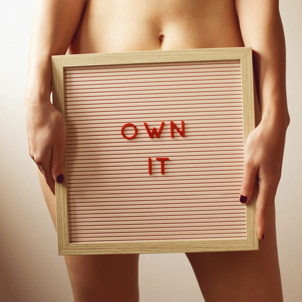 Frau hält Schild vor ihrem Schoßraum mit der Aufschrift „Own it“, Symbol für Selbstbestimmung und weibliche Autonomie